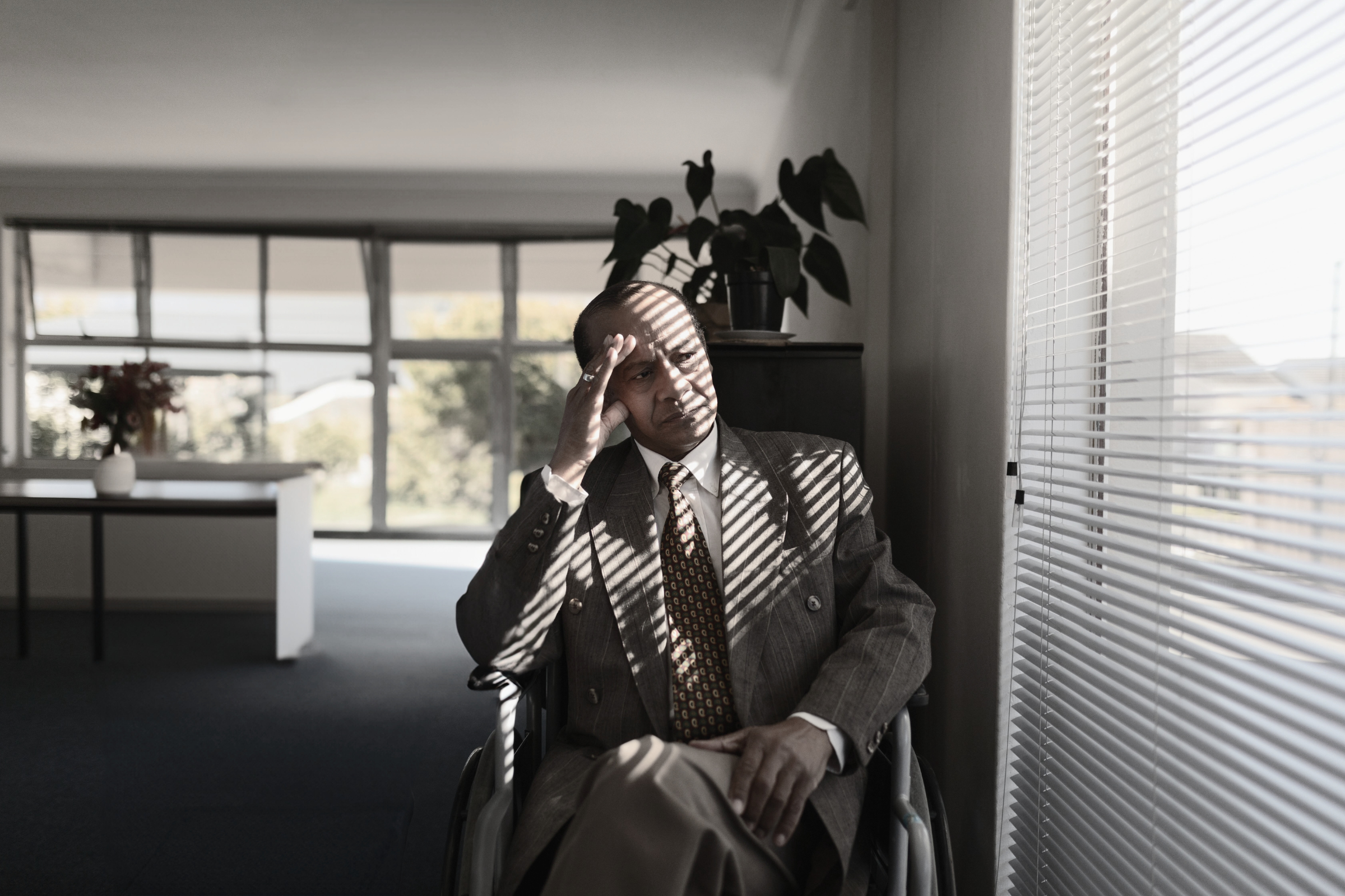 Businessman sitting in wheelchair near blinds casting striped shadows on suit in office lobby
